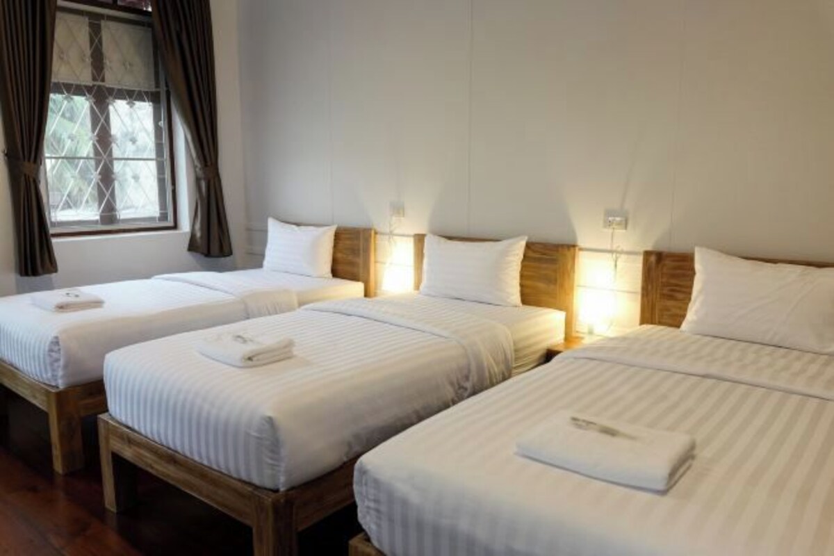 A hotel room with three single beds, each with a wooden headboard and white bedding. There is a window with curtains on the left side.