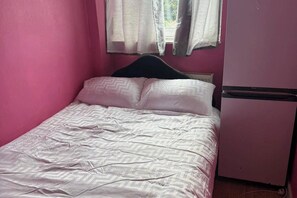 Uma cama de solteiro com colcha estampada, cabeceira e um pequeno frigorífico num quarto com paredes cor-de-rosa.