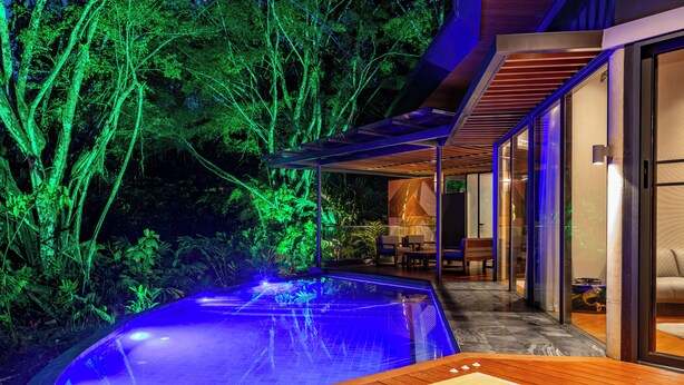 Un área de piscina moderna al aire libre con un deck de madera, puertas de vidrio que dan a un espacio de descanso y un exuberante fondo verde.