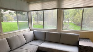 Un rincón con asientos acolchados, un banco y una pequeña mesa, ventanas amplias con persianas y vistas a un paisaje verde.