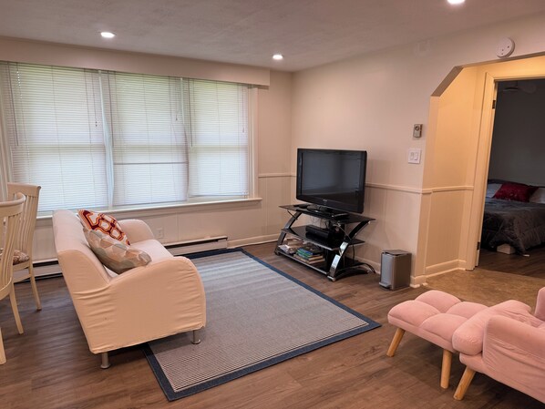 A living room with a sofa, a television on a stand, a small table, and a window with blinds.