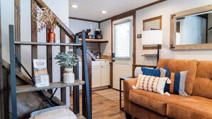 A cozy living area with a sofa, a bookshelf, a staircase, and a kitchen in the background. - Wayah Ridge Escapes (Topton)