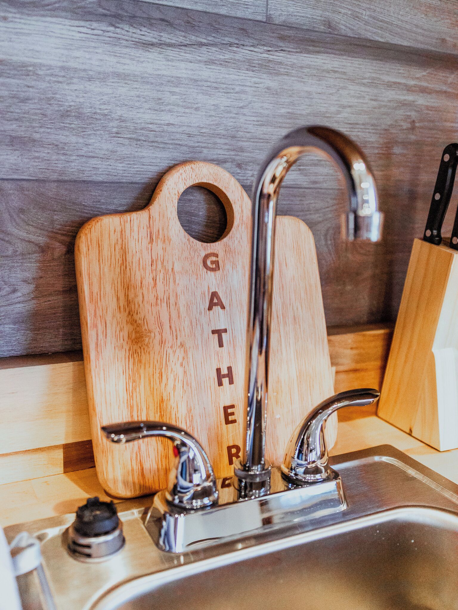 A kitchen sink with a wooden cutting board labeled "GATHER" and a knife block.
