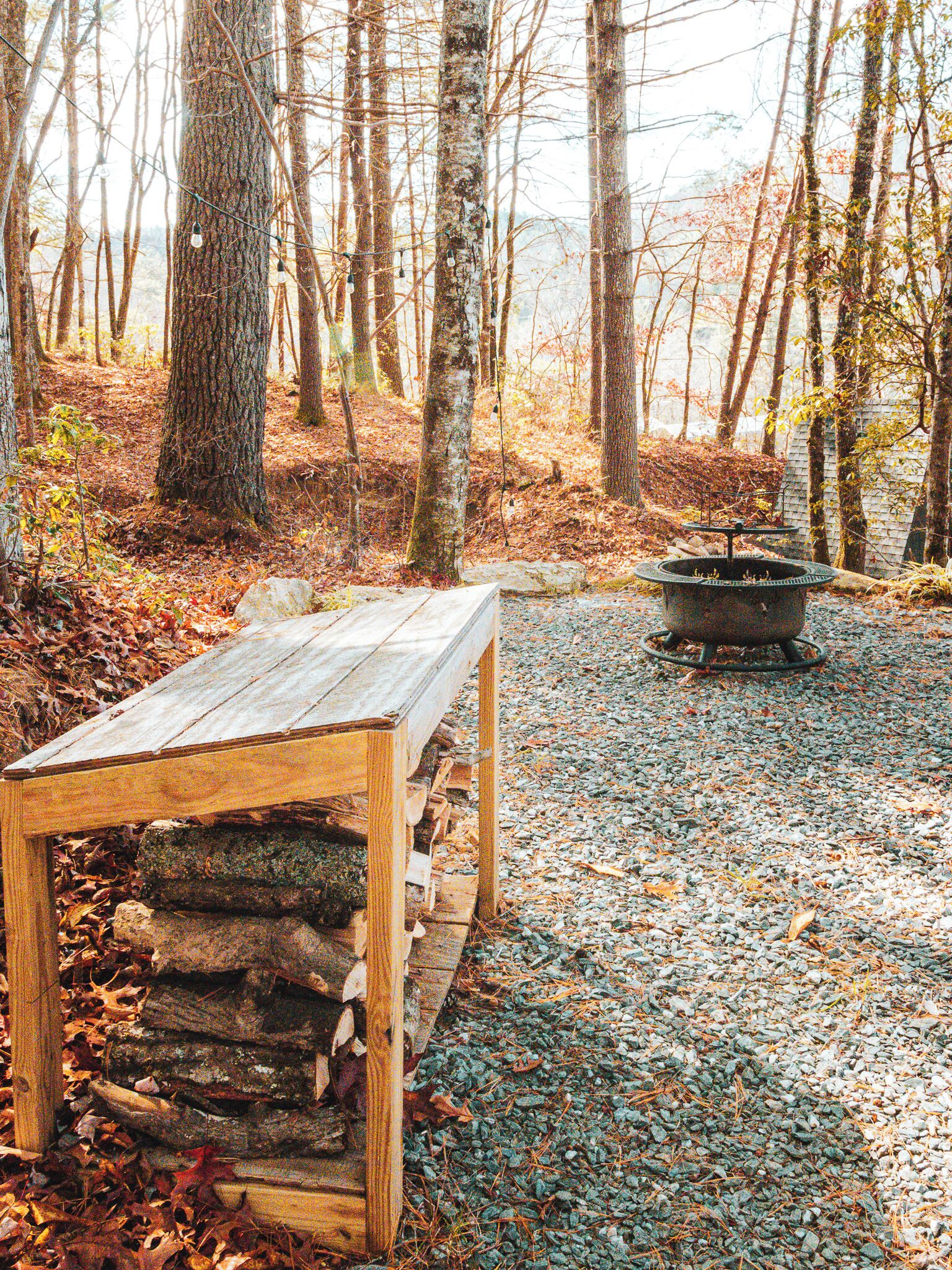 A wooden bench with a stone base in a forested area with a fire pit in the background.