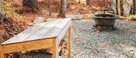 A wooden bench with a stone base in a forested area with a fire pit in the background.