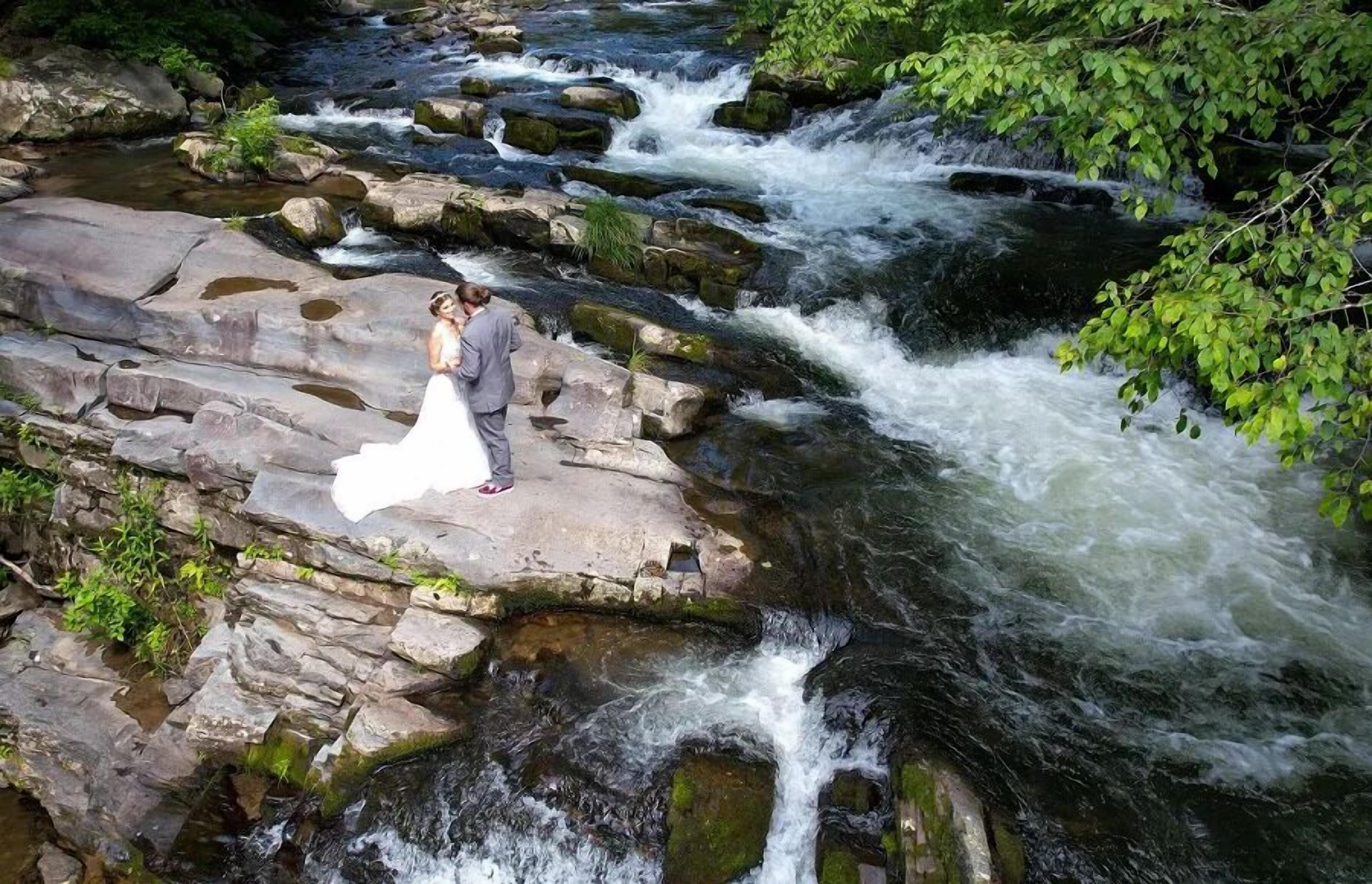 A couple standing on a rocky ledge by a flowing river.