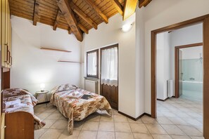 Un dormitorio con dos camas, techo de madera, una mesita de noche con lámpara y un baño visible a través de una puerta abierta.