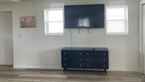 Habitación con televisor de pantalla plana en la pared, cómoda de color oscuro y dos ventanas con persianas.