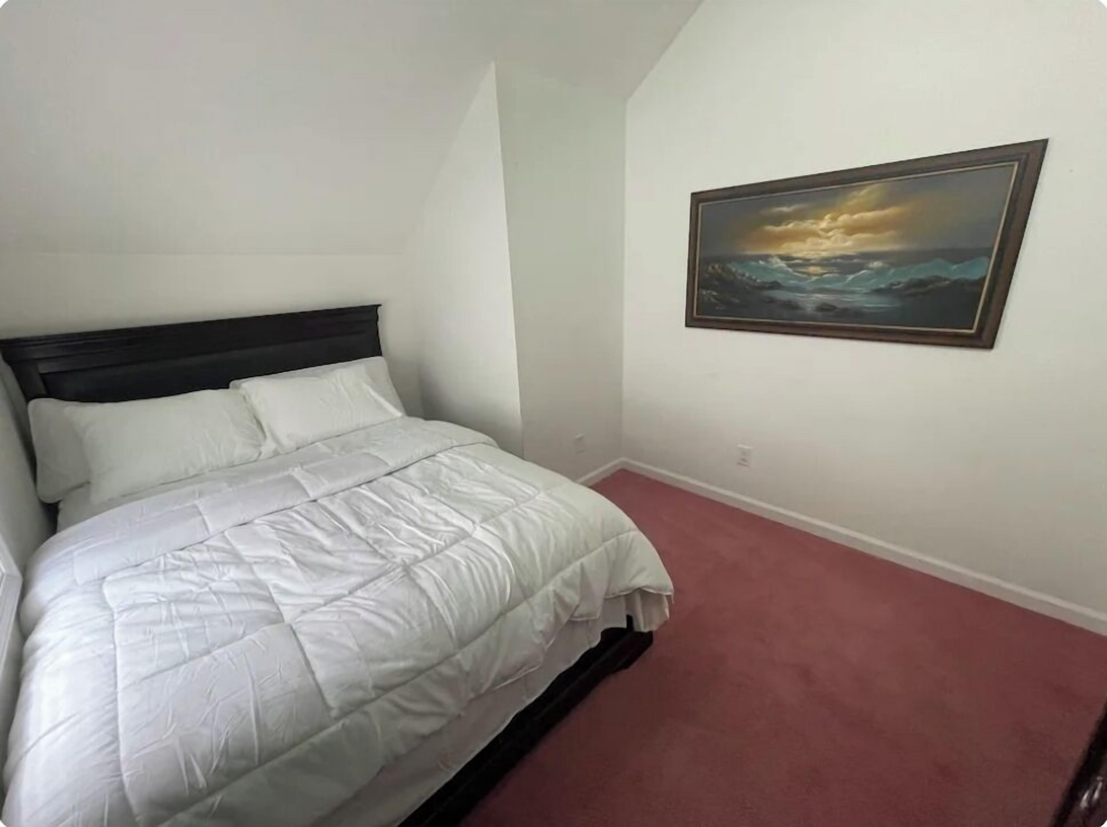 A bedroom with a bed, white bedding, a dark headboard, a painting of a seascape, and a red carpet.