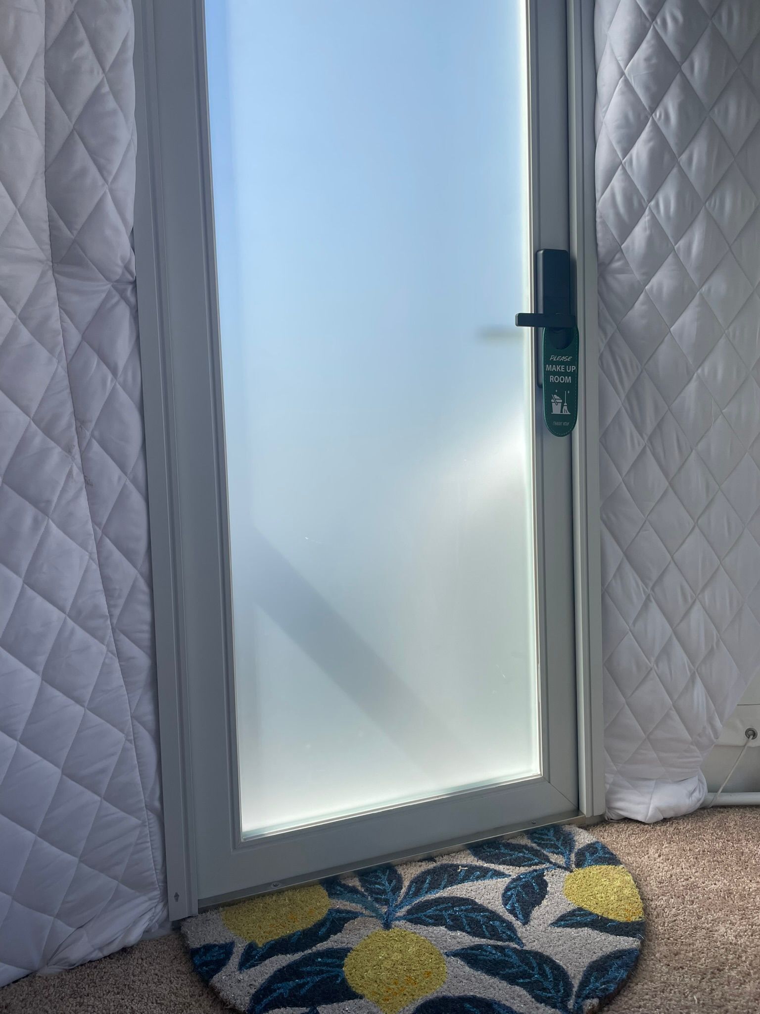 Une pièce avec une porte blanche matelassée, une porte en verre sur laquelle est apposée une pancarte indiquant « Veuillez préparer la chambre », et un tapis à motifs sur le sol.