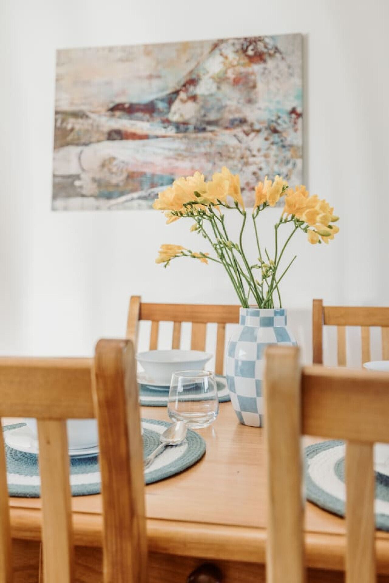 Ett matbord med en vas med gula blommor, trästolar och en rutig vas.