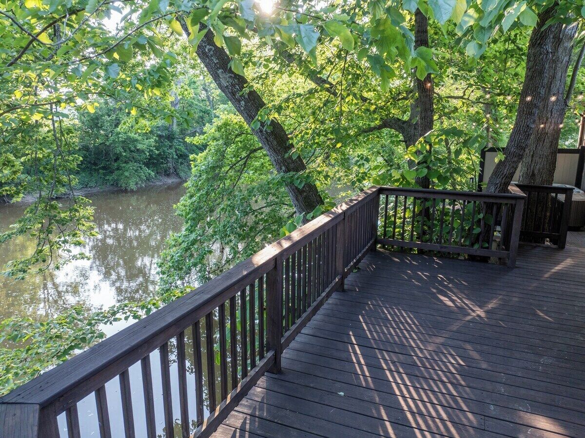 Une terrasse en bois dotée d’une balustrade, qui offre une vue sur une rivière paisible, le tout entouré d’une végétation luxuriante.