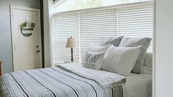 Un dormitorio con una cama, persianas blancas, una planta colgante y una lámpara.