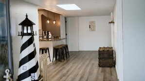 A room with a wooden floor, a small kitchen area with a coffee machine, stools, and a wall-mounted light fixture. - Pleasant Acres - Pleasant Acres (Greenville)