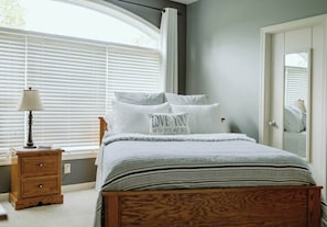 Une chambre à coucher comprenant un lit, une table de chevet, une lampe, une fenêtre avec des stores et une porte dotée d’un miroir.
