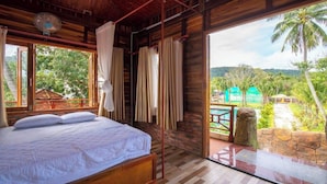 A room with a bed, wooden walls, a fan, and a view of a lush landscape. - New Serene (Phu Quoc)