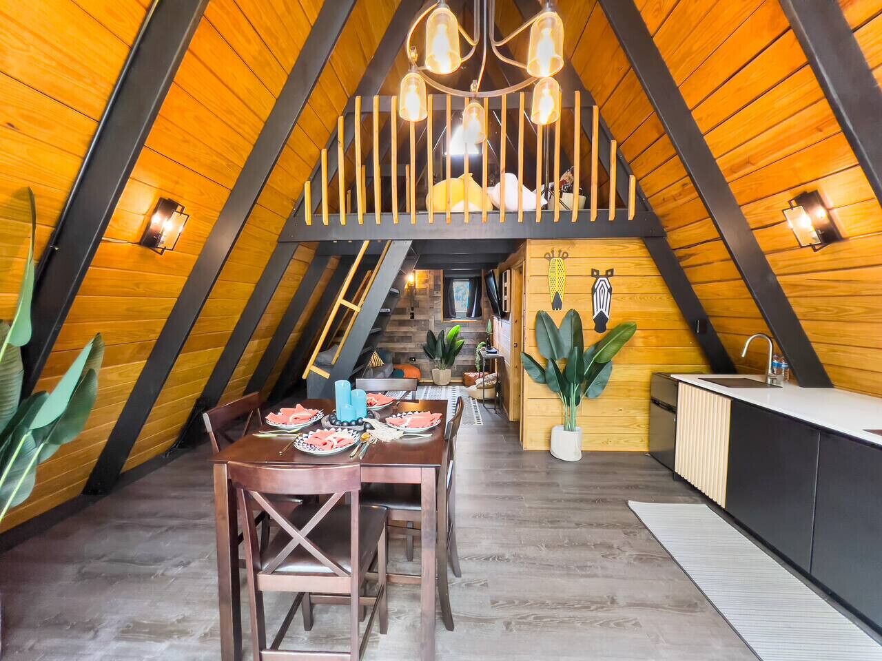 An attic-style room with a dining table set for four, a kitchen area with a sink and cabinets, and a staircase leading to an upper level.
