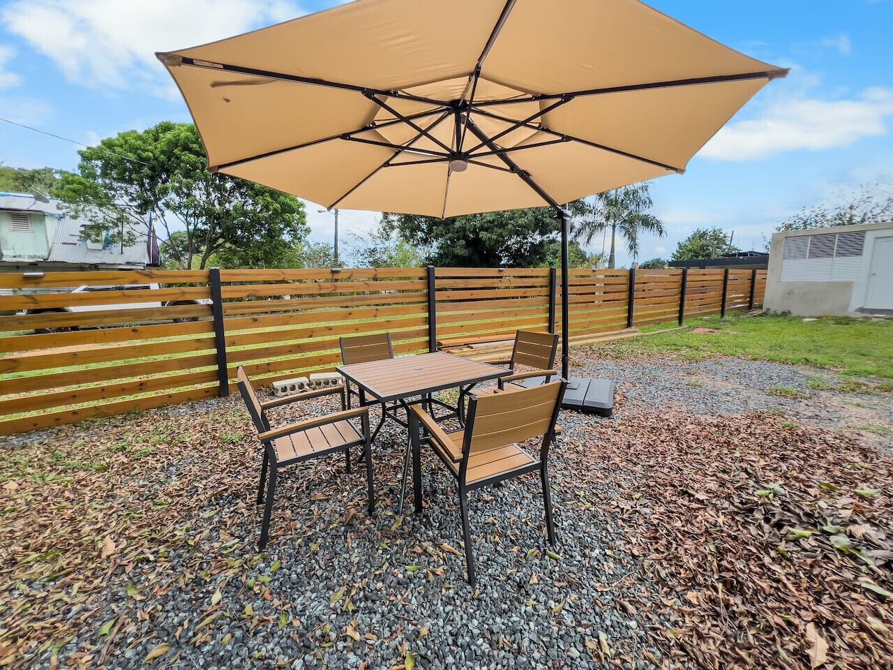 Une terrasse extérieure avec une table et des chaises sous un grand parasol, entourée d’une clôture en bois et d’un sol recouvert de gravier.