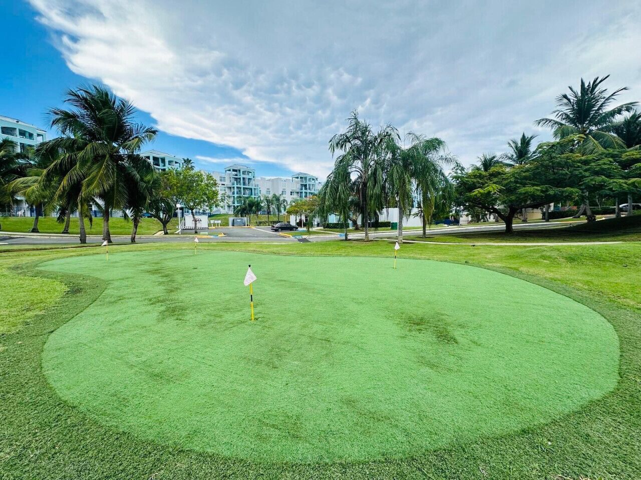 A golf course with a putting green, palm trees, and a building in the background.