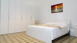 A bedroom with a bed, a nightstand, a lamp, a wardrobe, and a painting of a sunset over the ocean.