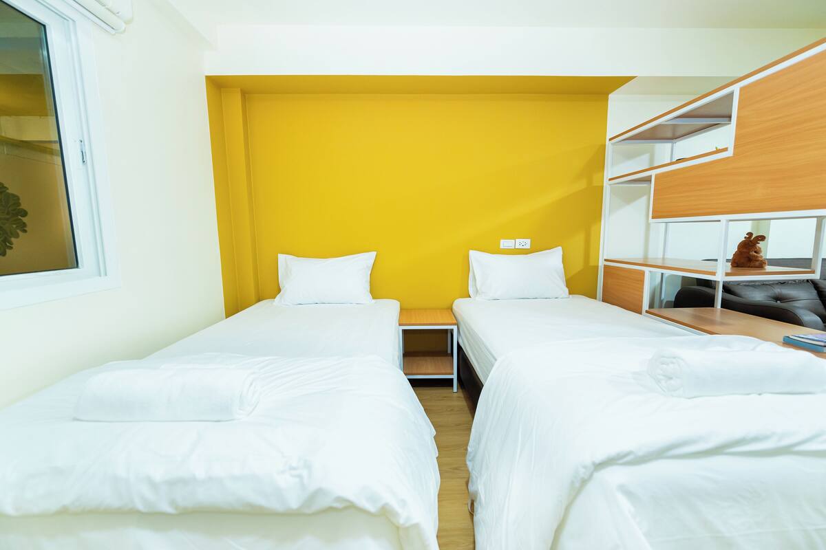 A hotel room with two single beds, a yellow accent wall, a small wooden table, and a built-in shelf with a toy.