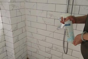 A person holding a spray bottle, likely for cleaning or disinfecting purposes, in a bathroom with white tiled walls and a visible shower area. - Julan'an Homestay (Guilin Liangjiang Sihu Dongxi Alley Shop) (Guilin)