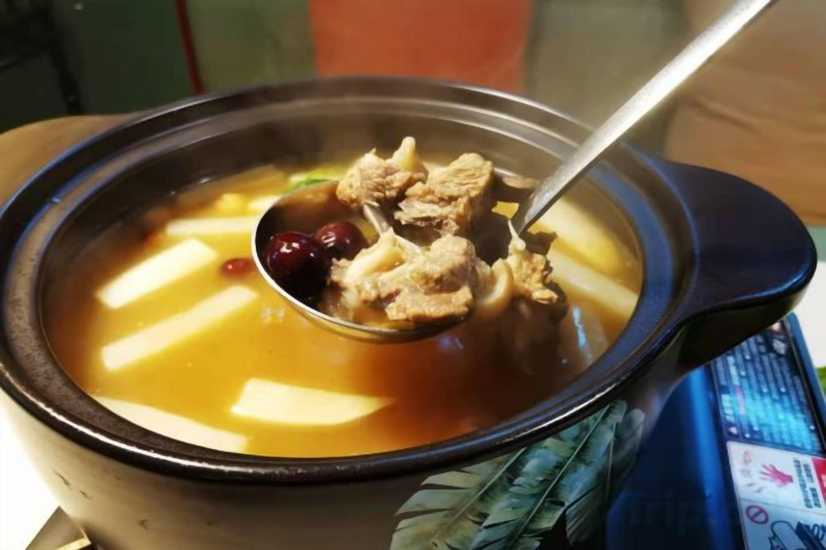 Un bol de sopa amb una cullera, una olla negra i un objecte blau a prop.