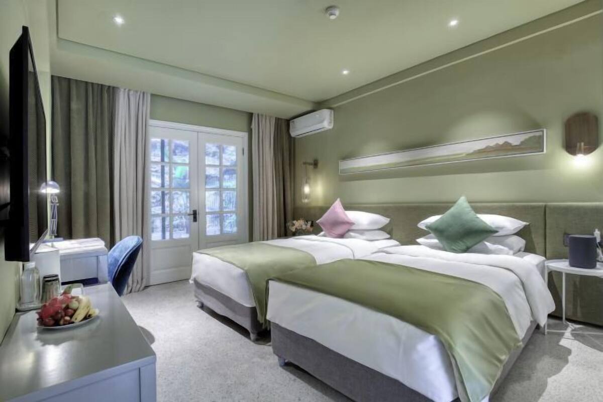 A hotel room with two beds, a desk with a lamp, a TV, and a window with curtains.