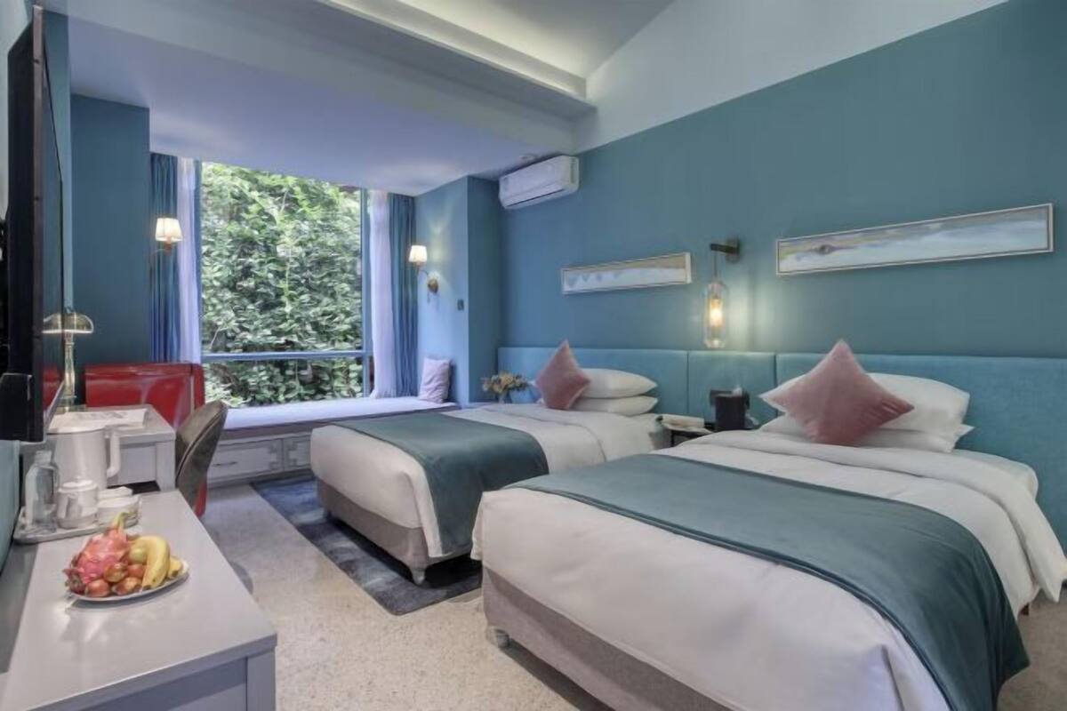 A hotel room with two beds, a desk with a computer, a window with a view of greenery, and an air conditioning unit.