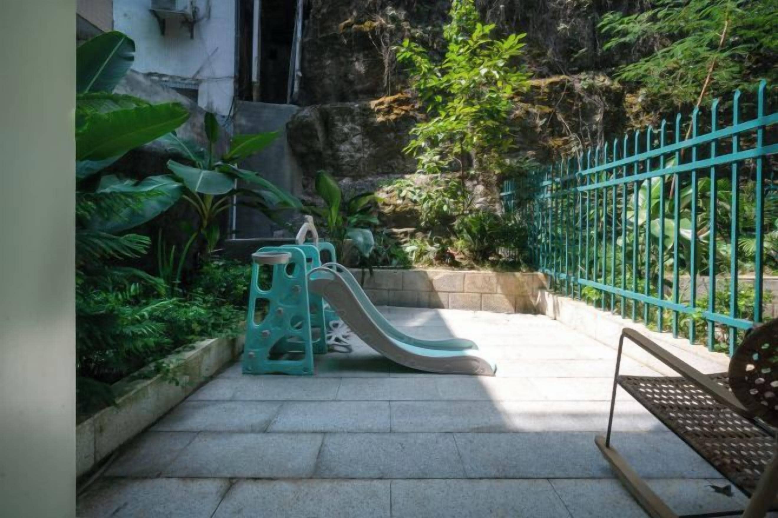 Une aire de jeux avec un toboggan et une structure d’escalade, entourée de verdure et d’une clôture bleue.