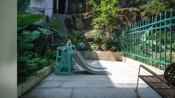 Une aire de jeux avec un toboggan et une structure d’escalade, entourée de verdure et d’une clôture bleue.