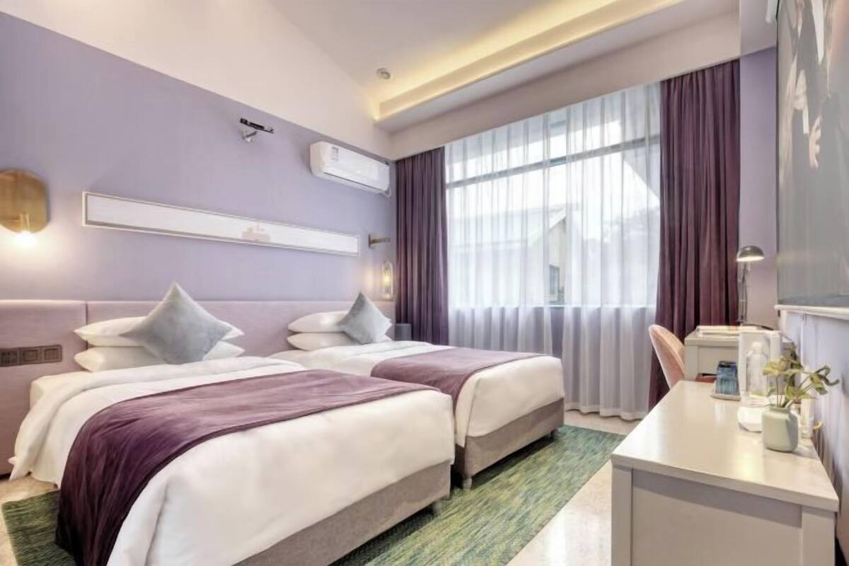 A hotel room with two beds, a desk, a chair, a window with curtains, an air conditioning unit, and a wall-mounted shelf.