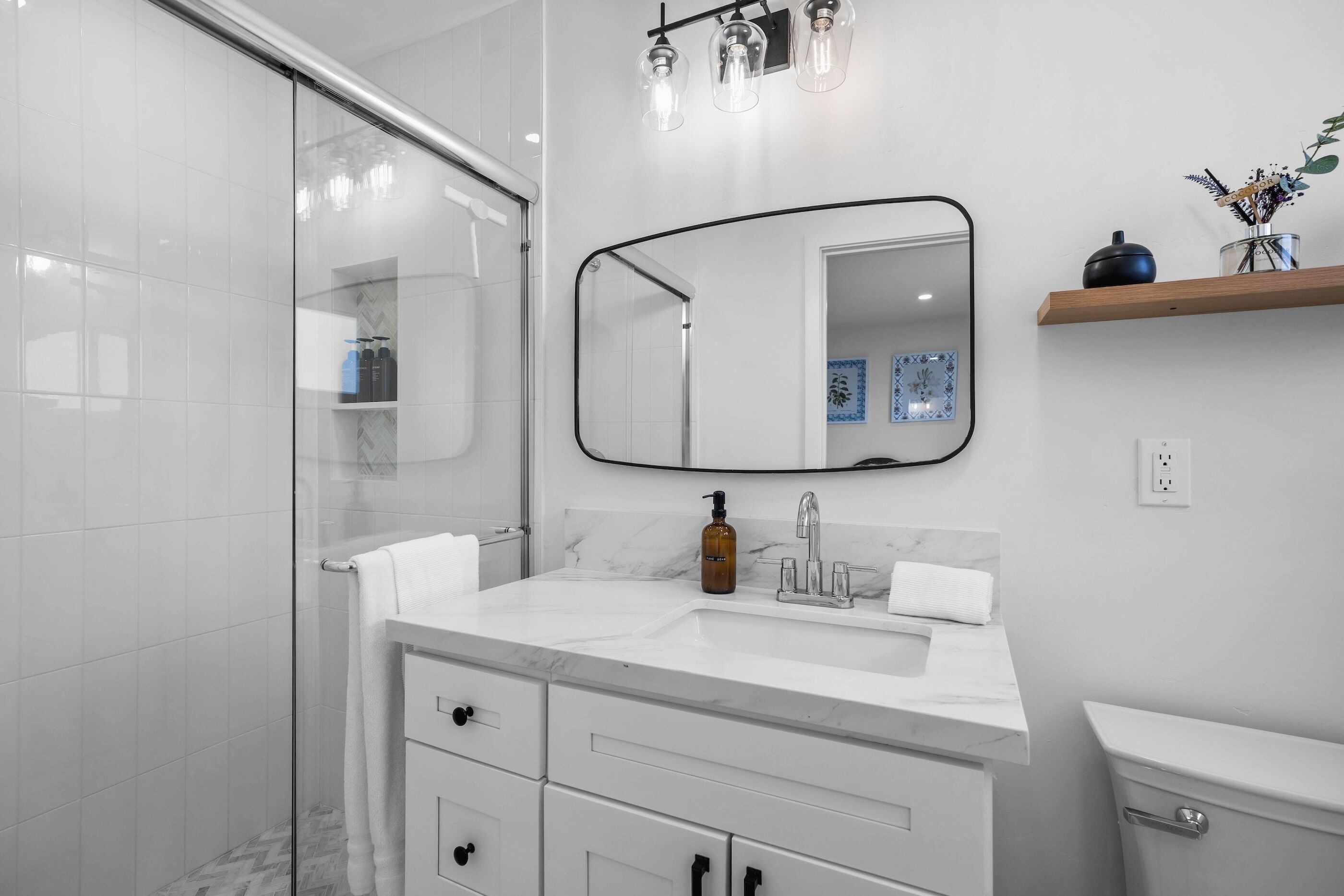 Une salle de bain moderne avec une douche à parois de verre, un meuble-lavabo blanc doté de tiroirs, un plan de toilette en marbre, un miroir avec un cadre noir et une petite étagère ornée d’objets décoratifs.