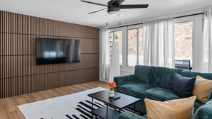 Ein modernes Wohnzimmer mit einem grünen Sofa, einem Flachbildfernseher, der an einer Wand aus Holzpaneelen befestigt ist, einem Deckenventilator und großen Fenstern mit weißen Vorhängen.