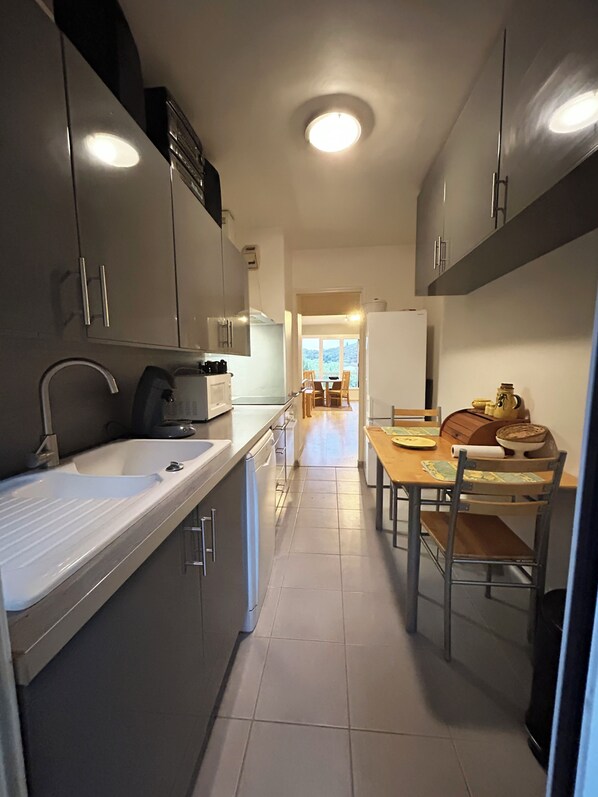 A narrow kitchen with a sink, stove, and cabinets. A dining area with a table and chairs is visible in the background. - Apartment with a garden in the Terres du Larzac and Lac du Salagou area (Lodève)