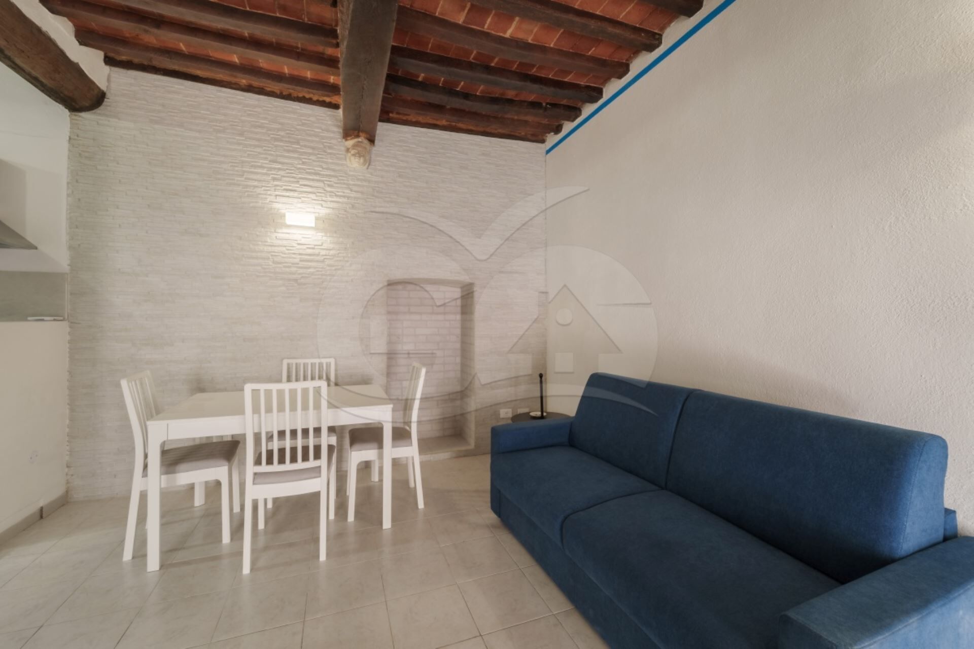 A room with a blue sofa, white chairs, a tiled floor, and a brick wall.