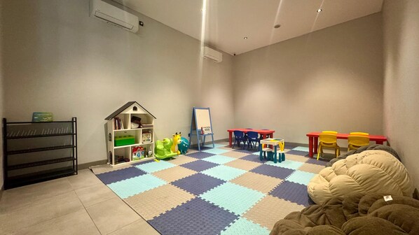 Un cuarto de juegos infantil con piso a cuadros, mesas de juego, sillas, una estantería y una pizarra.