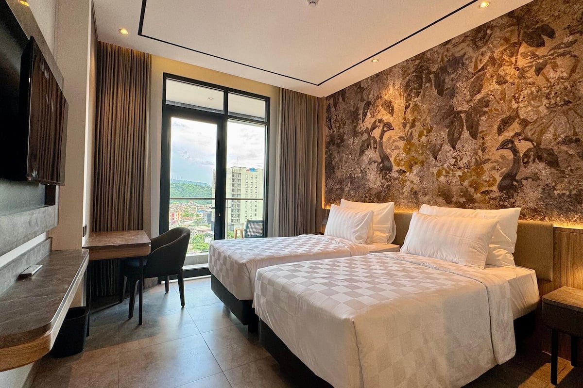 Een hotelkamer met twee bedden, een bureau, een stoel, een televisie en een groot raam met uitzicht op de stad.