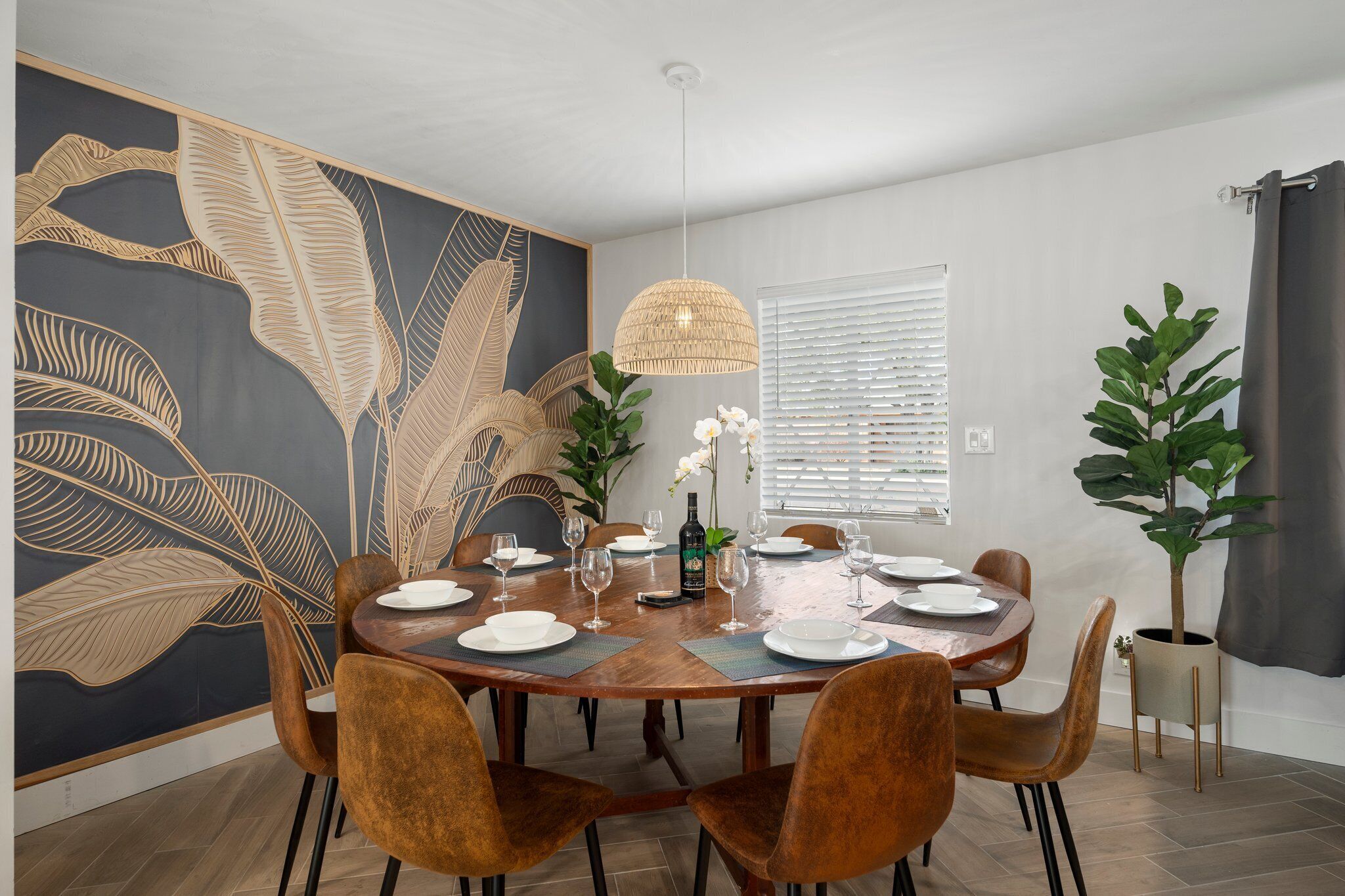 Ein Essbereich mit einem runden Holztisch, gedeckt für sechs Personen, umgeben von braunen Stühlen. An der Wand befindet sich ein großes Wandbild mit Blättermustern, eine Hängeleuchte und eine Zimmerpflanze stehen daneben.