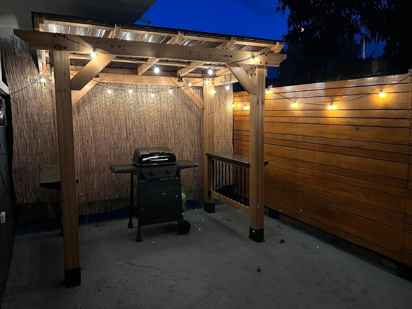 An outdoor patio area with a wooden pergola, a grill, and string lights.