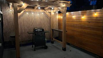 An outdoor patio area with a wooden pergola, a grill, and string lights.