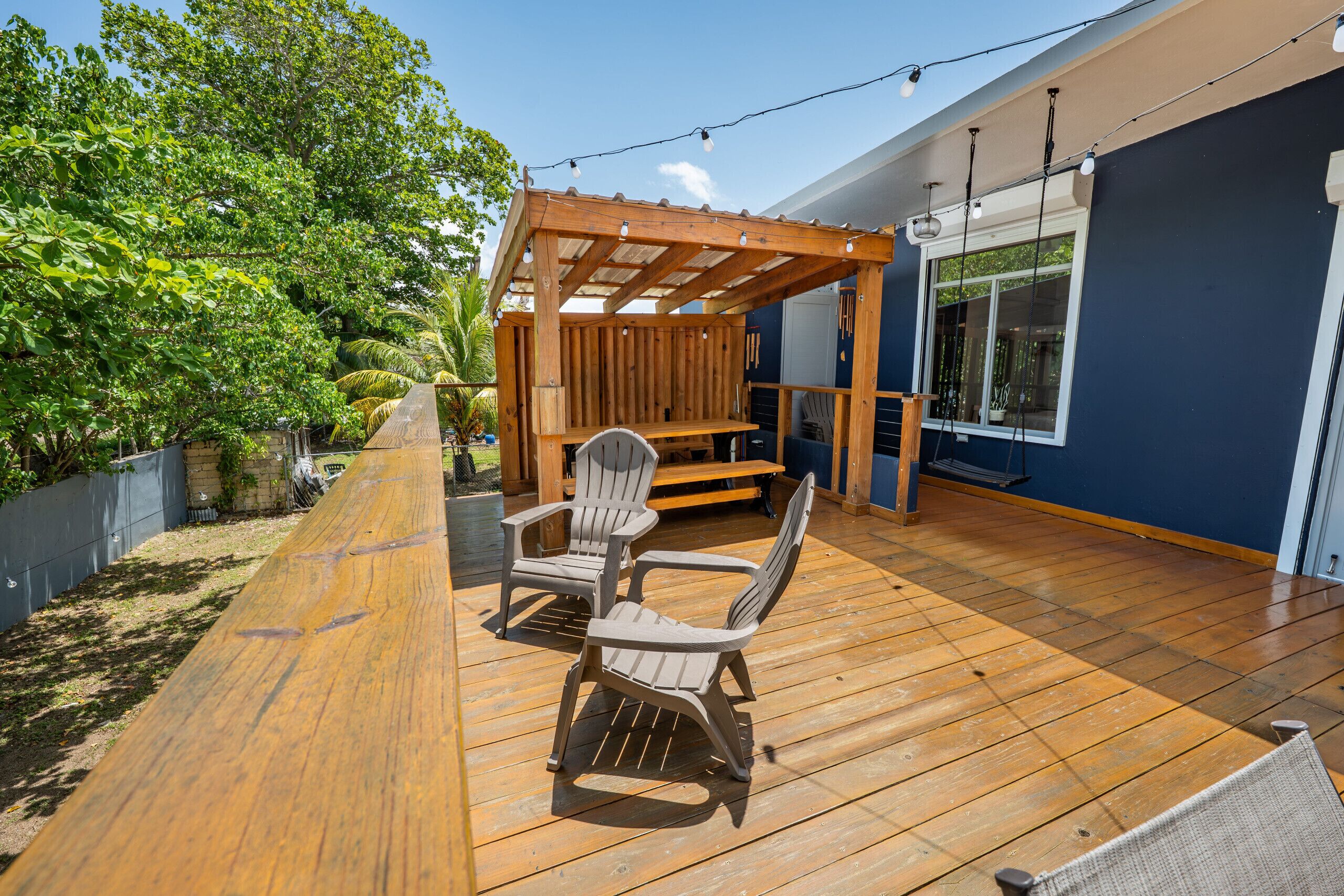 A wooden deck with Adirondack chairs and a pergola, surrounded by lush greenery.