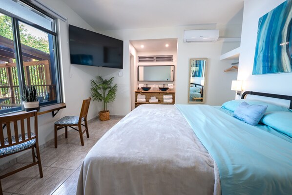 A hotel room with a large bed, a TV mounted on the wall, a bathroom with a mirror and sink, and a window with a view of greenery.