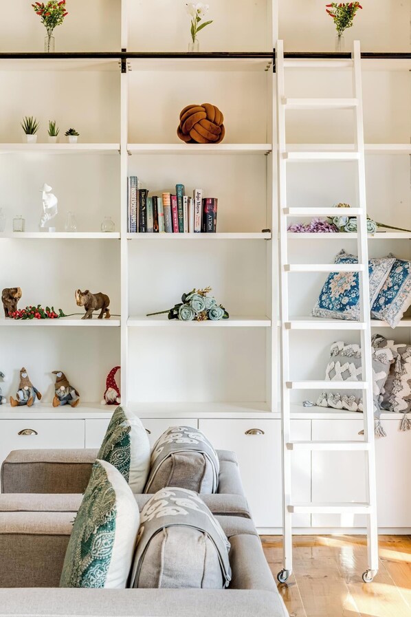 A modern living room with a grey sofa, a white ladder, and a bookshelf filled with books, decorative items, and a small plant.