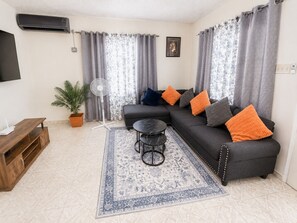 A living room with a sofa, a coffee table, a TV, a fan, and a potted plant.