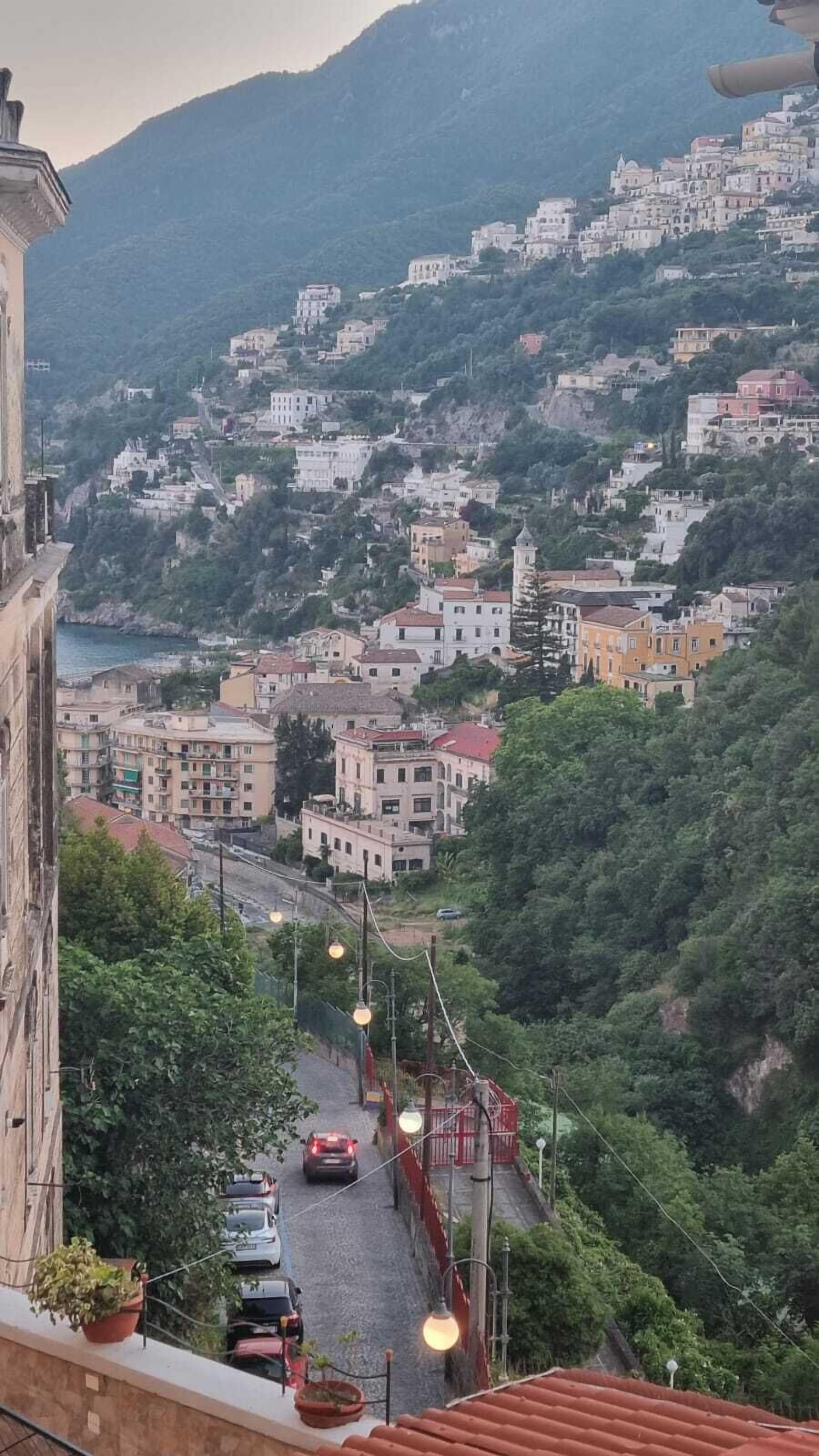 Un pueblo en una ladera con edificios, una carretera y una masa de agua a lo lejos.