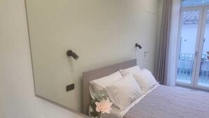 Habitación de hotel con un espejo grande, cama con ropa blanca, mesita de noche con un jarrón de flores y ventana con vistas al exterior.