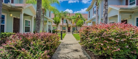 A pathway flanked by buildings with palm trees and shrubs, leading to a building with a visible door number.