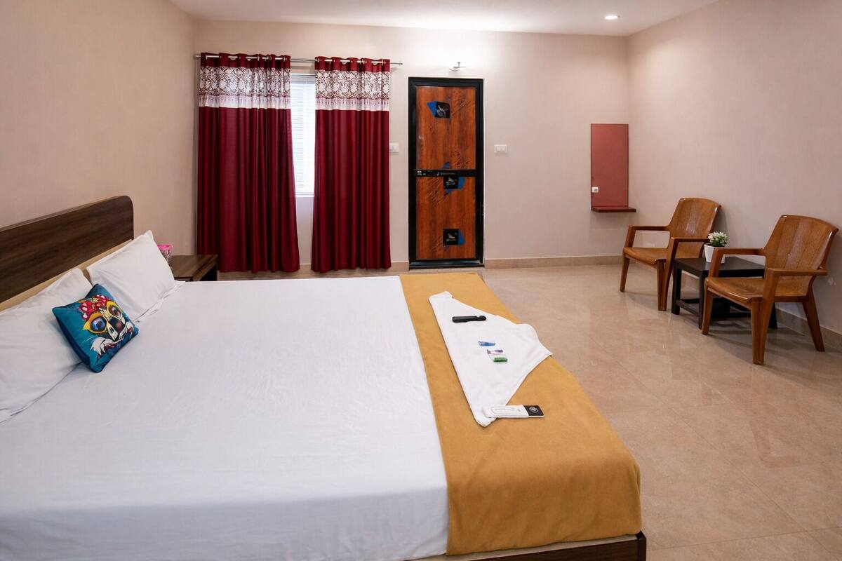 Une chambre d’hôtel avec un lit, un bureau, deux chaises, une porte et une fenêtre avec des rideaux.