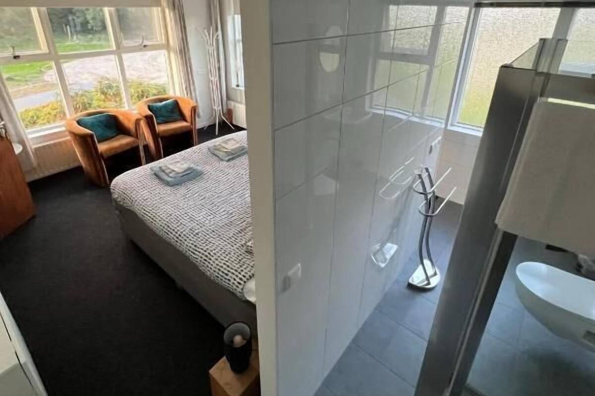 A modern hotel room with a large bed, a sitting area with a chair and table, a bathroom with a sink and mirror, and large windows offering a view of the outdoors.